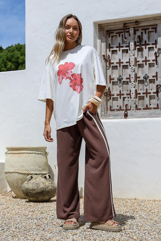 Woman wearing a white oversized t-shirt with pink floral design and brown pants standing in front of a white wall.