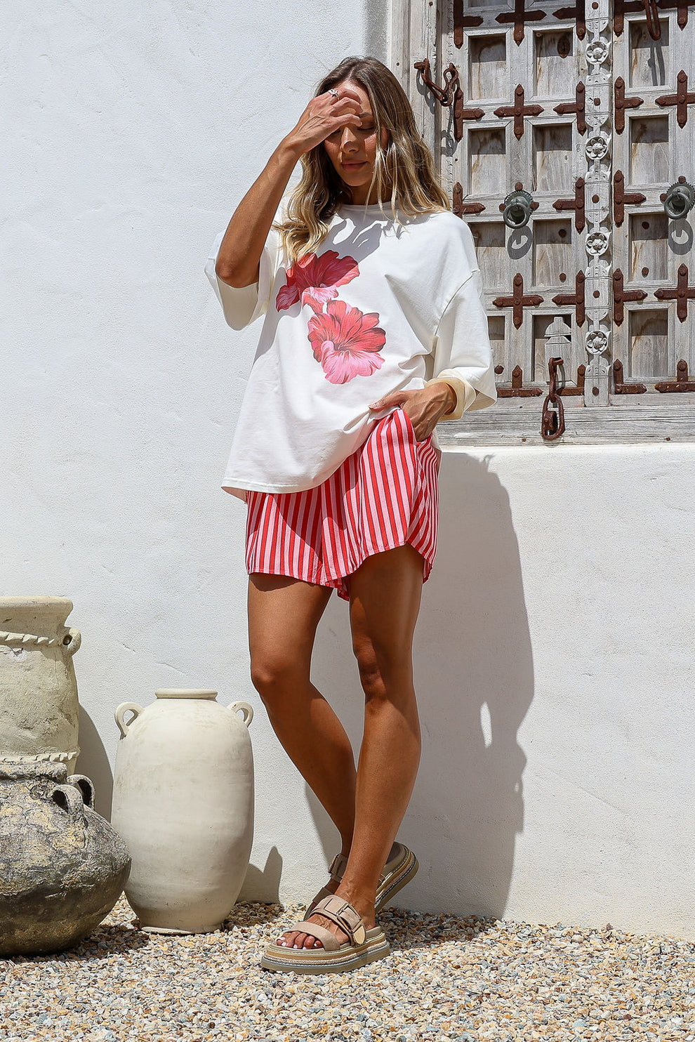 Woman wearing a white oversized shirt with pink floral design and red and white striped shorts, standing against a white wall with decorative elements.